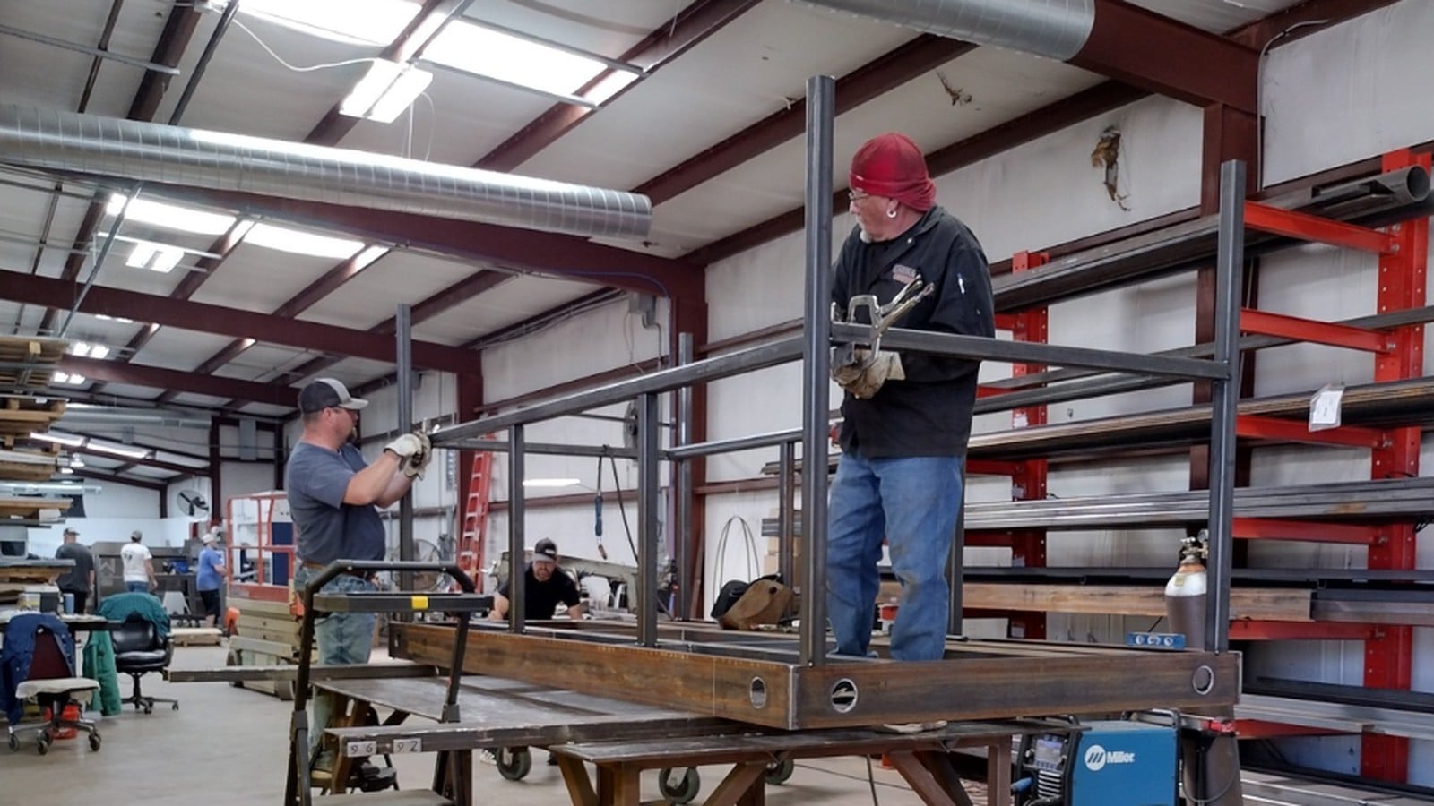 Two men build a large steel frame, one standing on the frame while the other holds a crossbar in place.
